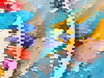 Brushes on a palette showing a variety of paint colours. The palette is over a canvas of an impressionistic painting of multi-coloured brush strokes.