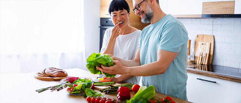 Happy couple in modern kitchen preparing healthy meal together with fresh colorful vegetables including lettuce, peppers, asparagus, and tomatoes on counter, demonstrating nutritious cooking for brain health, mental wellness, and cognitive function.