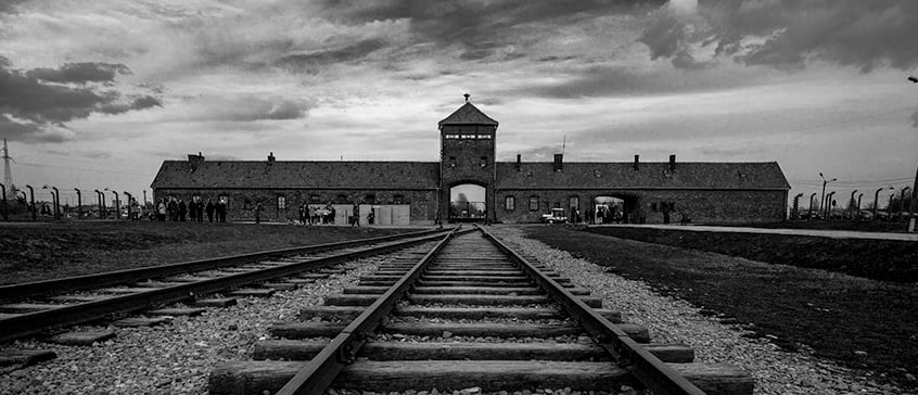 Black-and-white photo of railway tracks leading to the entrance of Auschwitz concentration camp under a cloudy sky, symbolizing historical memory and dark tourism.