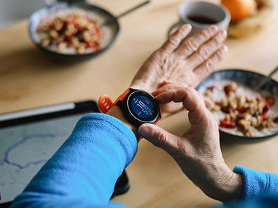 Close-up of a smartwatch displaying health data next to a tablet with a map and bowls of food, illustrating age-tech tools for healthy, independent living.