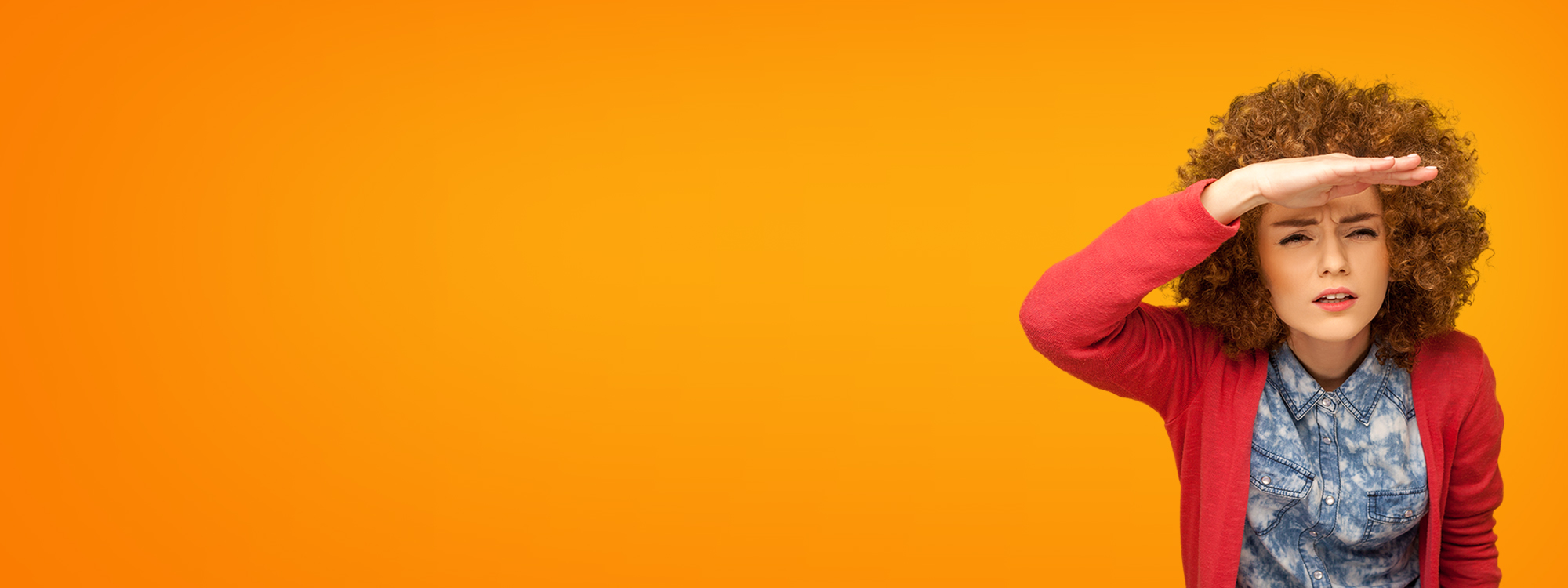 Curly-haired woman in red sweater and denim shirt shielding her eyes, looking ahead against orange background.