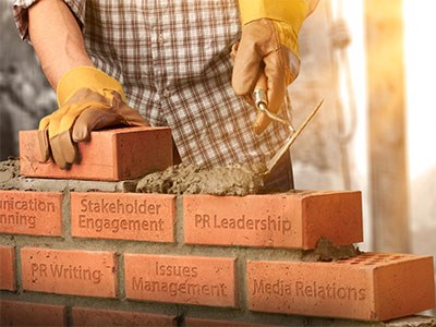 Construction worker layering bricks which have the following phrases engraved on them: stakeholder engagement, PR leadership, PR writing, issues management and media relations.
