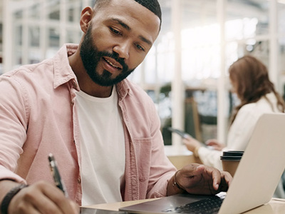 Man casually and smartly dressed concentrates on a writing task at his worktable. As we works at his laptop he writes notes on a note pad.