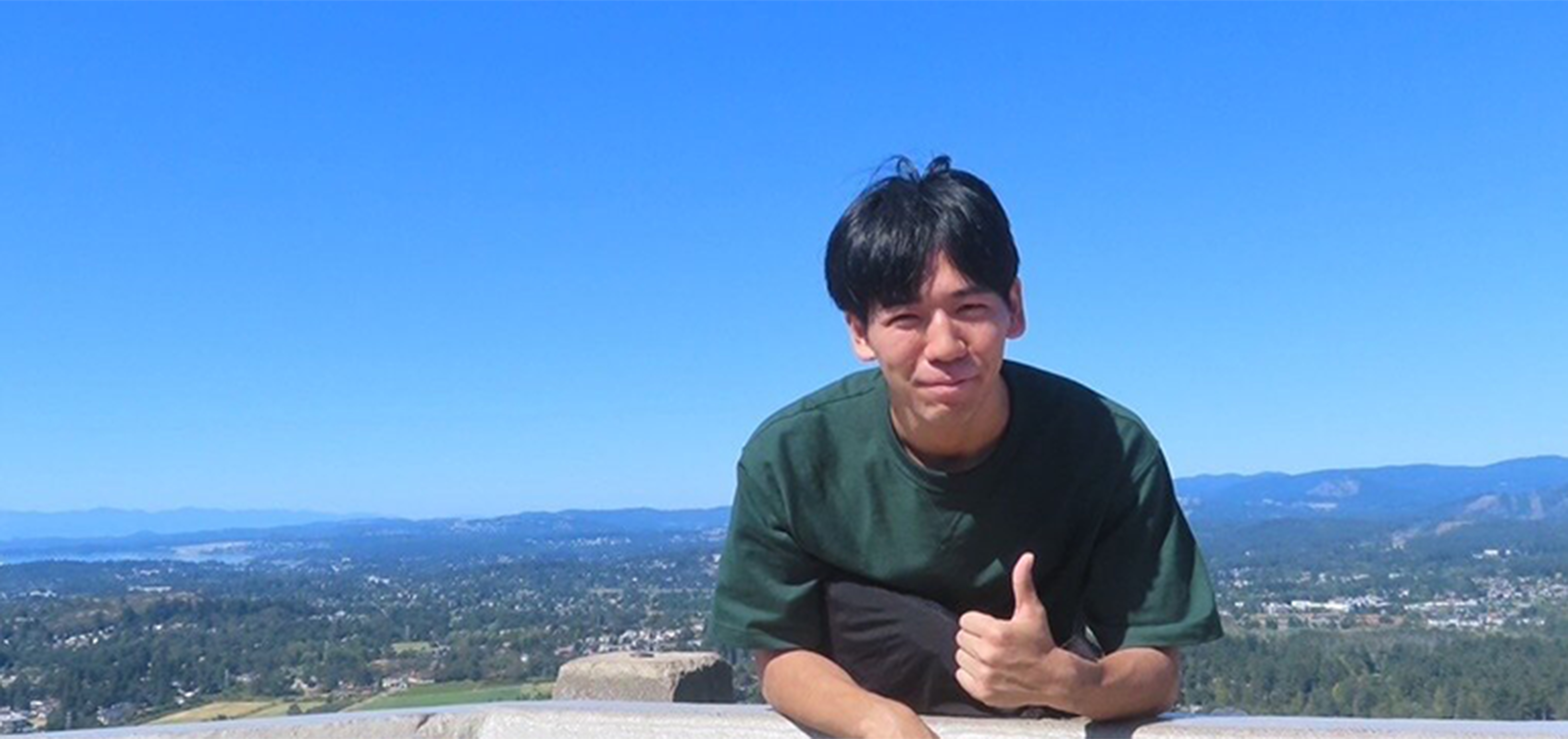 ELC student Akihito Koyama giving a thumbs up with a scenic view of Victoria and the surrounding landscape behind him.