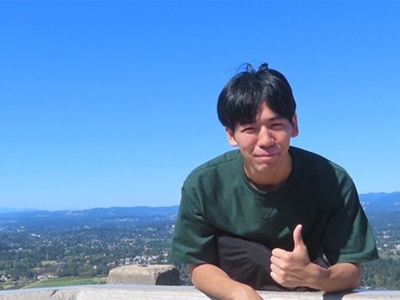 ELC student Akihito Koyama giving a thumbs up with a scenic view of Victoria and the surrounding landscape behind him.