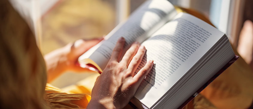 Close-up of hands holding an open book with sunlight casting shadows on the pages, symbolizing the immersive experience of reading and literary appreciation.