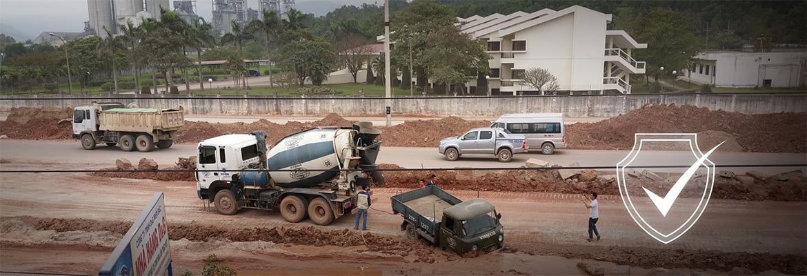 Construction site with concrete mixer trucks and dump trucks on dusty, unpaved ground. Workers visible near heavy machinery without clear protective equipment. Residential buildings nearby suggest potential community exposure to construction dust and noise pollution, impacting both worker respiratory health and air quality for local residents.
