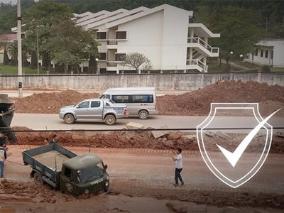 Construction site with concrete mixer trucks and dump trucks on dusty, unpaved ground. Workers visible near heavy machinery without clear protective equipment. Residential buildings nearby suggest potential community exposure to construction dust and noise pollution, impacting both worker respiratory health and air quality for local residents.