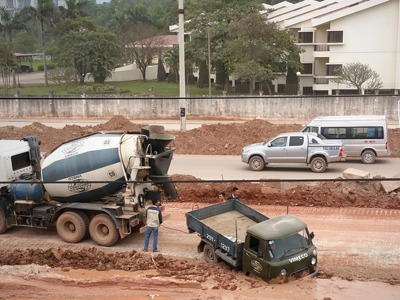 Construction site with concrete mixer trucks and dump trucks on dusty, unpaved ground. Workers visible near heavy machinery without clear protective equipment. Residential buildings nearby suggest potential community exposure to construction dust and noise pollution, impacting both worker respiratory health and air quality for local residents.