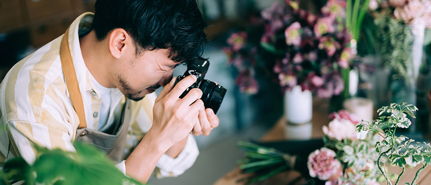 Photographer with glasses capturing close-up shots of plants in bright indoor space filled with potted flowers and greenery, demonstrating creative composition techniques in natural light