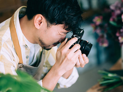 Photographer with glasses capturing close-up shots of plants in bright indoor space filled with potted flowers and greenery, demonstrating creative composition techniques in natural light