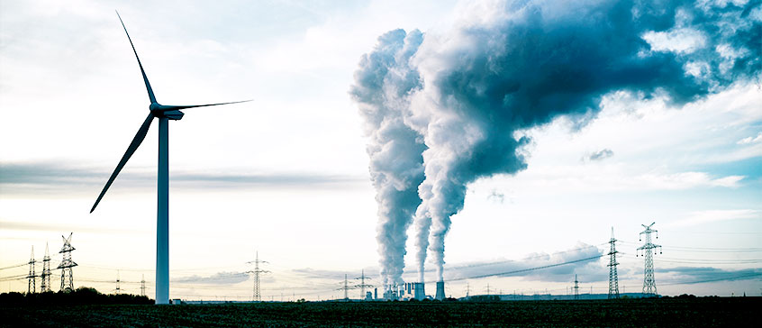 Photo of a green energy windmill juxtaposed with industry smoke towers. 