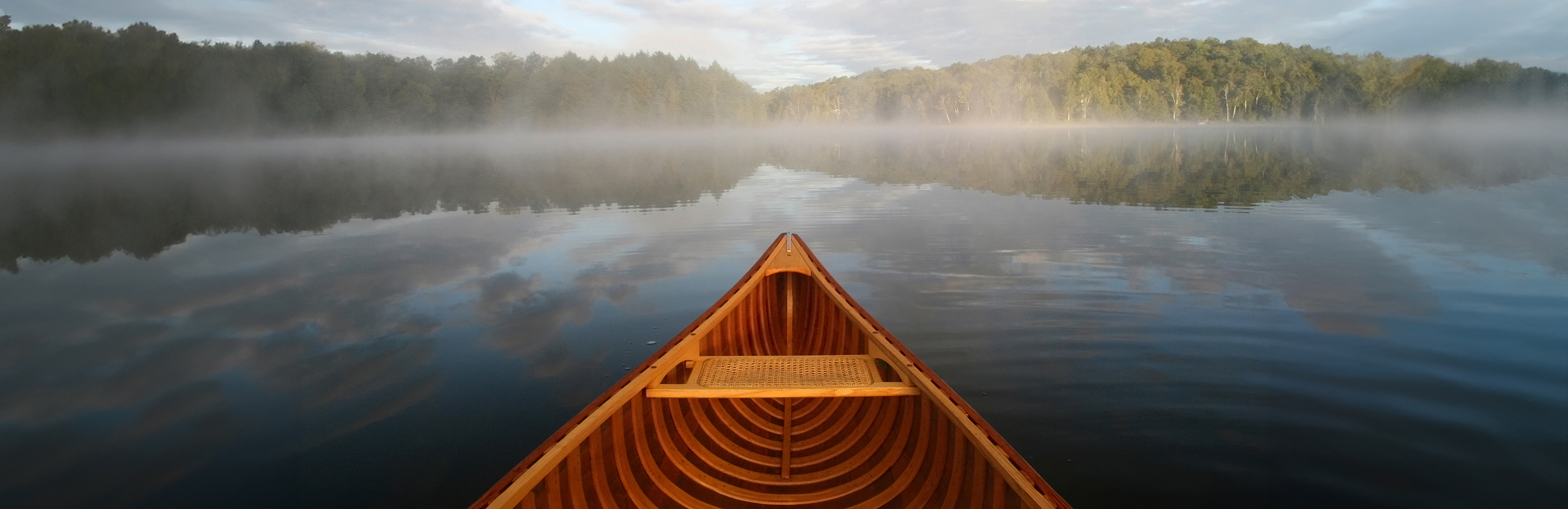 View from a canoe. The prow is visible as you look out over a calm lake to the tree-lined shore. Morning mist lightly hovers over the lake in bright morning sun.