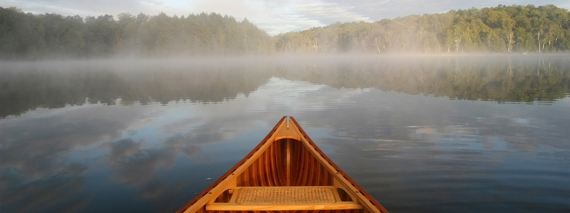 The bow of a wooden canoe on a calm, misty lake surrounded by trees at dawn.