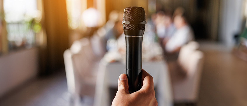 Close-up of a hand holding a black microphone toward the camera, with blurred people sitting in the background, representing the act of gathering personal stories and testimonials to craft meaningful tributes about someone