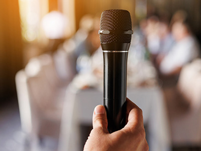 Close-up of a hand holding a black microphone toward the camera, with blurred people sitting in the background, representing the act of gathering personal stories and testimonials to craft meaningful tributes about someone