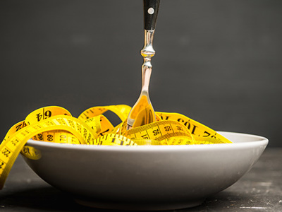 A gray ceramic bowl is filled with a coiled yellow measuring tape, with a silver fork stuck upright into the center of the tape. The measuring tape