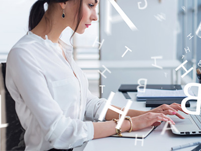 Woman wearing a white blouse with sleeves rolled up is sitting in profile typing at her laptop. Her dark hair in a ponytail. As she types white letters float around her.