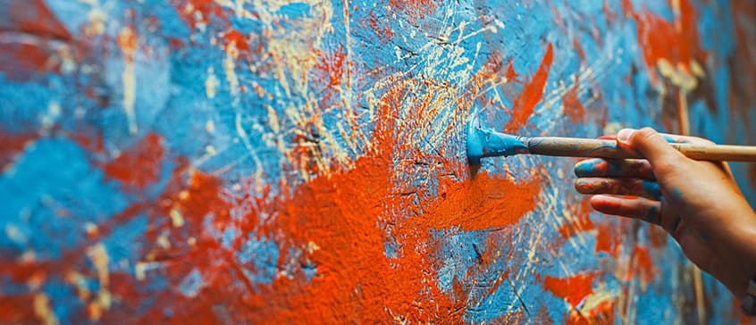 Close-up of hands holding paintbrushes creating an energetic abstract painting with vibrant orange and blue paint splatters and brushstrokes on canvas, demonstrating dynamic painting techniques and colour application.