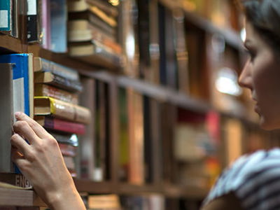 Books arranged on shelves in a library with a hand reaching for a blue-covered book, symbolizing exploration of Indigenous culture through literature.