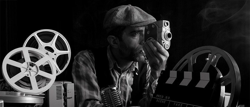 Black and white image of man in flat cap using vintage camera surrounded by film reels and clapperboard, evoking classic filmmaking and experimental cinema techniques.