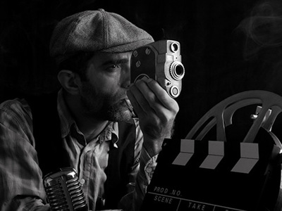 Black and white image of man in flat cap using vintage camera surrounded by film reels and clapperboard, evoking classic filmmaking and experimental cinema techniques.