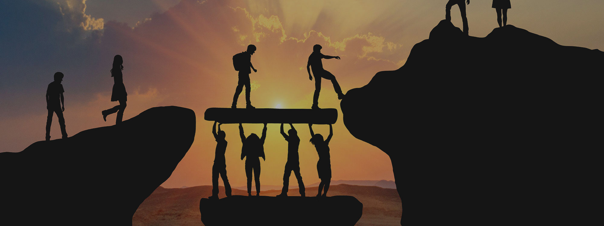 Silhouette of people on a mountain at sunset, helping each other climb and celebrating atop a peak.