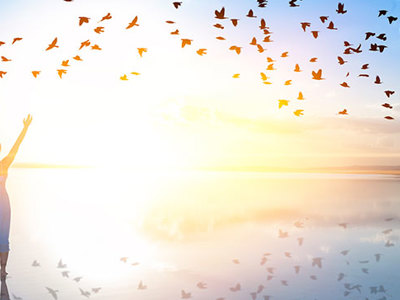 A person standing on a beach with arms raised towards a flock of birds flying across a sunrise.