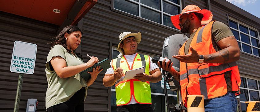 Three individuals wearing safety vests and hard hats standing outside a building, holding clipboards and devices, engaged in discussion about safety procedures and emergency planning.
