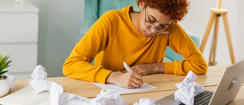 A person with curly red hair and glasses wearing a yellow sweater sits at a wooden desk, focused on writing by hand on paper. Crumpled papers are scattered around the workspace, with a laptop open nearby, illustrating the creative writing process and revision work.