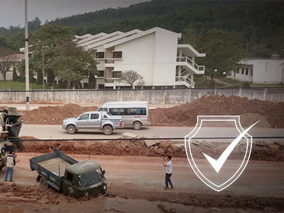 Construction site with concrete mixer trucks and dump trucks on dusty, unpaved ground. Workers visible near heavy machinery without clear protective equipment. Residential buildings nearby suggest potential community exposure to construction dust and noise pollution, impacting both worker respiratory health and air quality for local residents.