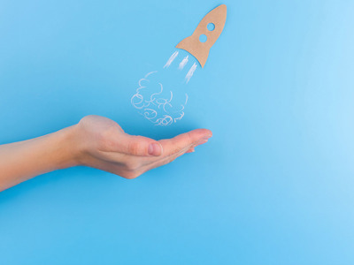 A hand beneath a sketched rocket ship taking off against a light blue background. 