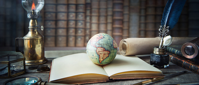 Vintage study scene with an open book, a small globe, an ink bottle with a quill, rolled parchment, a magnifying glass and an oil lamp on a wooden desk, set against shelves of old books, symbolizing history and global exploration.