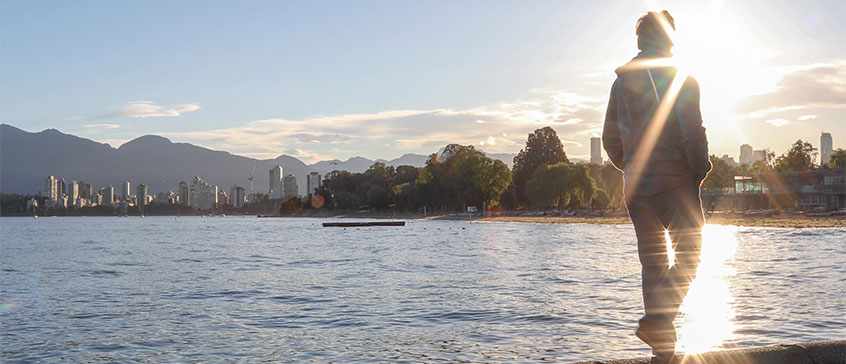 Person walking along a body of water with an urban skyline on the other side of the water.  