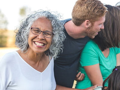 A diverse group of people of various ages, including an older woman with curly gray hair and glasses in the foreground, gathered together outdoors in a warm, social setting. The group appears relaxed and engaged in friendly conversation, illustrating healthy social connections and balanced interpersonal relationships.