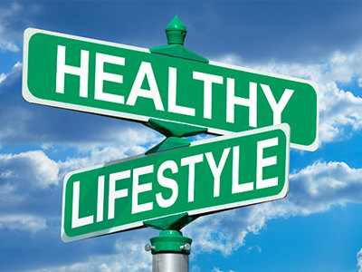 A street signpost with two green directional signs against a bright blue sky with white clouds. The top sign points left and reads HEALTHY in white capital letters. The bottom sign points right and reads LIFESTYLE in white capital letters. The image symbolizes choices and directions toward a healthier life