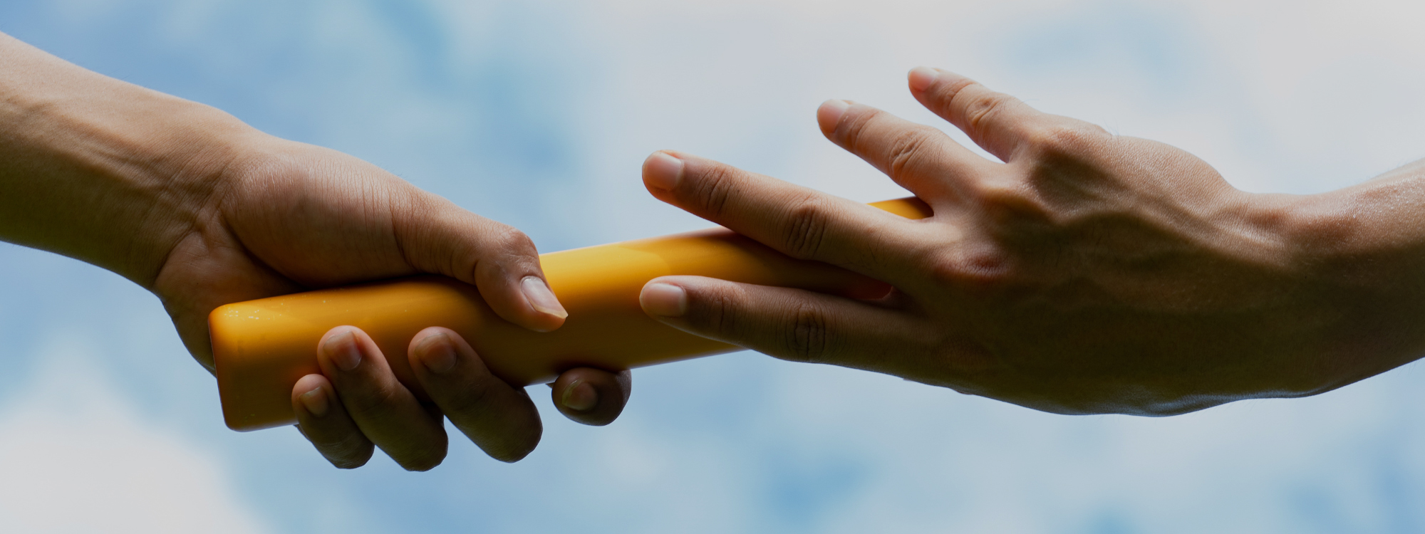 Close-up of two hands passing a bright yellow relay baton against a blue sky with soft white clouds.