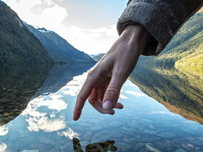 A hand gently touching the surface of clear lake water, creating small ripples. The water reflects the sky, mountains, and a few clouds. The lakebed is visible through the clear water, covered with small rocks. Green mountains rise on either side of the lake under a bright sky.