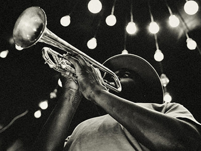 Black and white photograph of musician playing trumpet under string lights, capturing the intimate atmosphere and musical heritage central to blues origins and development